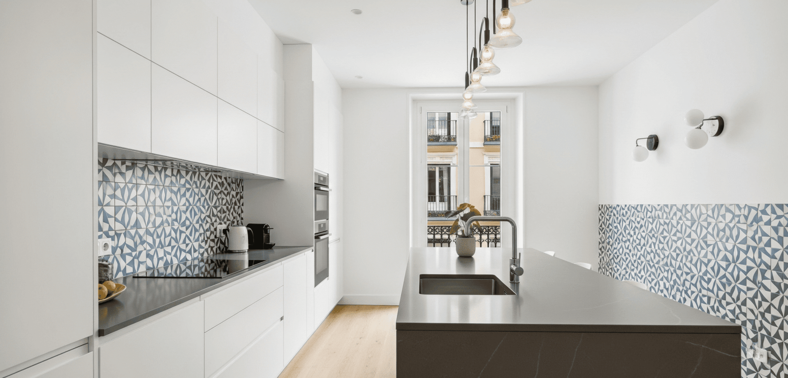 Cocina moderna con isla central resultado de una reforma de cocina en Madrid, destacando el mobiliario blanco sin tiradores y los azulejos geométricos.